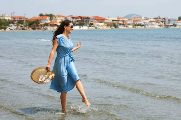 青いドレスを着た若いブルネットの女性がビーチを裸足で歩き 水の中で足をぶら下げています 夏休み明けましておめでとう — ストック写真