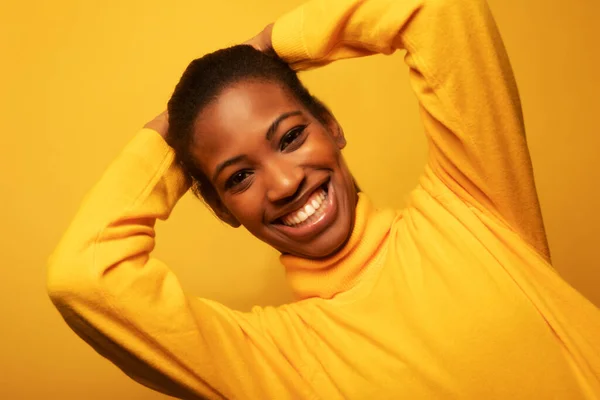 Attractive African American woman in a yellow sweater smiles happily with her arms crossed behind her head. Portrait on a yellow studio background.