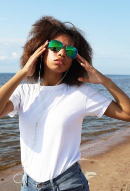 Güneş gözlüklü, kulaklıklı, cep telefonuyla müzik dinleyen büyüleyici afro-amerikalı genç kadın. Sıradan giyinmiş. Denize yakın bir yerde eğlenmek için. Yaşam biçimi.