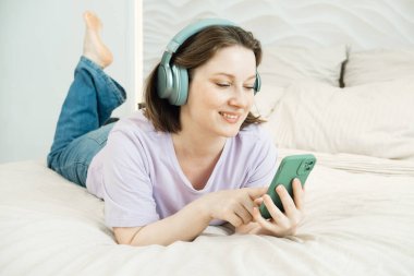 Gülümseyen yetişkin kadın kulaklıklarla yatağa uzanıyor ve elinde akıllı bir telefonla raylara bakarken müzik dinliyor.