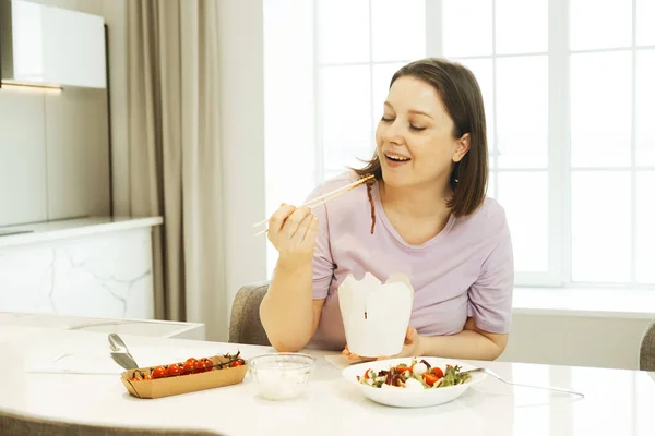 かなり豊満な女が台所に座り 紙の白い箱から麺を食べる 食料の配達だ 不健康な食べ物 — ストック写真