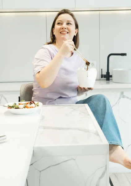 漂亮的胖女人坐在厨房里 吃着纸盒里的面条 食品交付 不健康的食物 — 图库照片