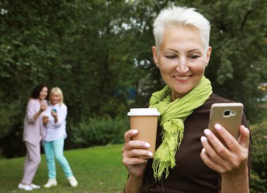 An elderly blond lady takes a selfie, holding a cup of coffee to go, in the background her female friends also use smartphones. Sunny day in the summer park. Golden Age of Retirement.
