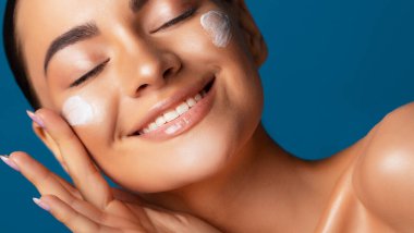 Close up beauty portrait of a beautiful half naked woman applying face cream with closed eyes over blue background