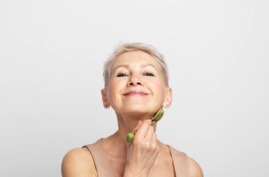 Beauty face portrait of cute and pretty senior woman with short hair using facial roller massager for perfect fresh skin on her cheek, isolated on gray background