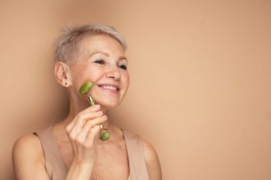 Gorgeous woman of 60s massaging face skin using natural facial stone trendy jade green quartz roller device for detox of perfect skin. Spa technique skincare ads.