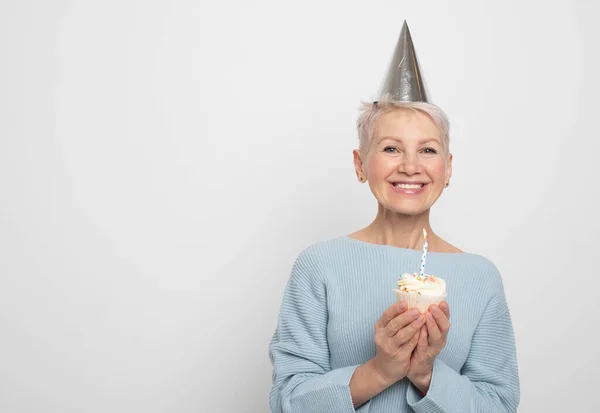 美しいです高齢者女性身に着けています円錐帽子お祝いの誕生日とともにカップケーキとともにキャンドルグレーの背景 — ストック写真