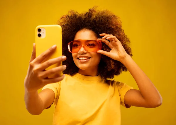 Young dark skinned mulatto woman with Afro hairstyle, holds mobile phone, makes video call, dressed in casual wear and orange sunglasses, isolated over yellow studio wall.
