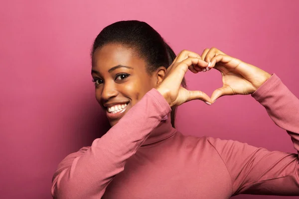 Young beautiful African American afro woman smiling in love doing heart symbol shape with hands over pink color background. Romantic concept.