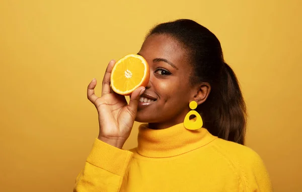 lifestyle, food, diet and people concept: Optimist Young female model of afro appearance. Clean, even skin of the face. In the hands of citrus, orange. Photo on yellow background.