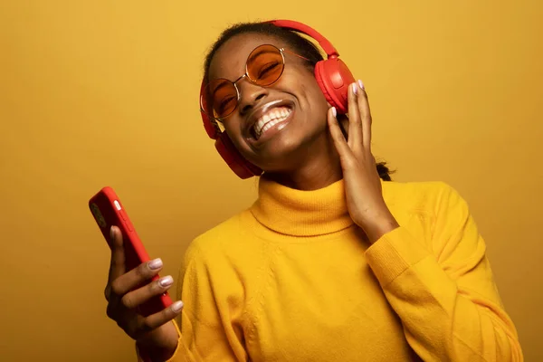 People, music, emotions concept. Delighted carefree afro american female with dances in rhythm of melody, closes eyes listens loud song in headphones, holds smart phone has fun.