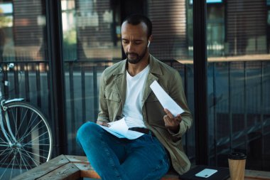 A young African American man sits at a bus stop and reads documents. Lifextyle concept.