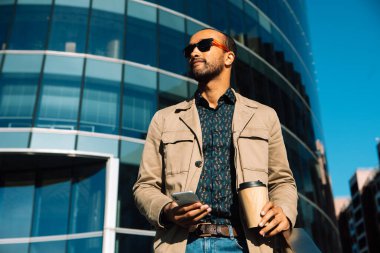 Young afro Man texting on phone. Casual urban professional entrepreneur using smartphone smiling happy outside office building. Outdoor portrait of modern young guy with mobile in the street