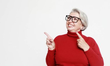 Smiling senior gray-haired woman wearing eyeglasses and red sweater isolated on grey studio background pointing with finger at blank copy space aside