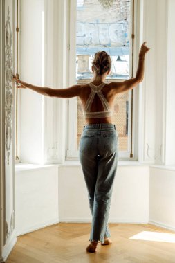 young blond woman in jeans and a lace top is standing in the living room, near big window, back view