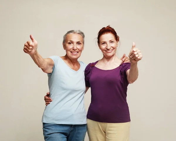 Two pretty older women friends show thunbs up on grey background. Lifestyle concept.