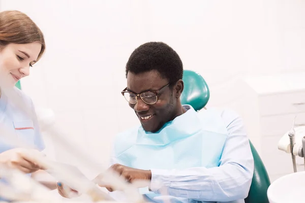 若い女性歯科医は黒人男性の治療計画を立てる 医者はタブレットを使う 生活習慣 医療の概念 — ストック写真