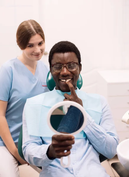 People, medicine, stomatology, technology and health care concept. Young smiling afro man looking in the mirror after dental procedure.