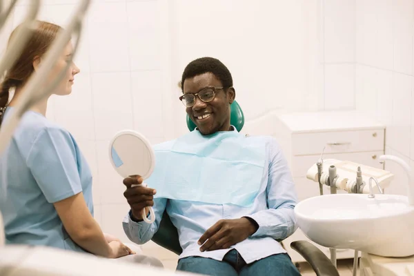 People, medicine, stomatology, technology and health care concept. Young smiling afro man looking in the mirror after dental procedure.