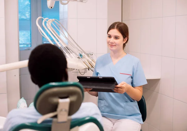 A young woman dentist makes a treatment plan for a black man. The doctor uses a tablet. People, medicine, stomatology, technology and health care concept.