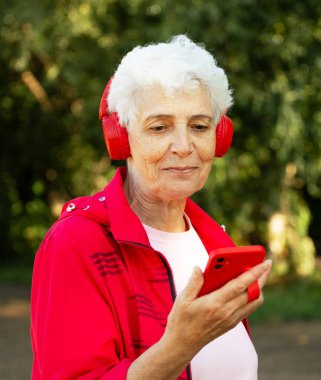 An elderly woman with a short haircut dressed in red sportswear listens to music on headphones, using a smartphone while jogging. Summer time.