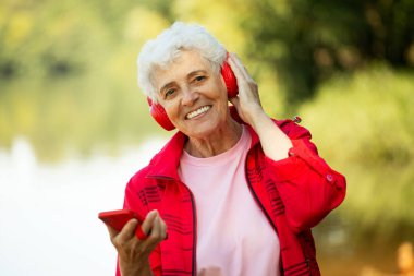 An elderly woman with a short haircut dressed in red sportswear listens to music on headphones, using a smartphone while jogging. Summer time.