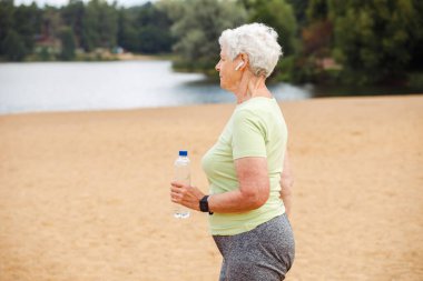 Elderly happy retired woman jogging. An old woman runs along the beach and holds a bottle of water. Summer time.