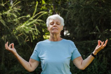 Mature woman relaxing in nature. Old woman meditating. Senior woman is doing yoga in the park. Smiling aged lady exercising. Old female workout. Sport training. Rehabilitation. Elderly woman.