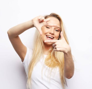 Body language, emotions, and people concept: Young blonde woman smiles and looks through a frame of fingers over white background