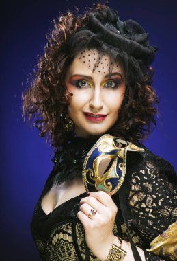 Beautiful curly woman with bright makeup and hat with veil holding venetian mask over blue color background