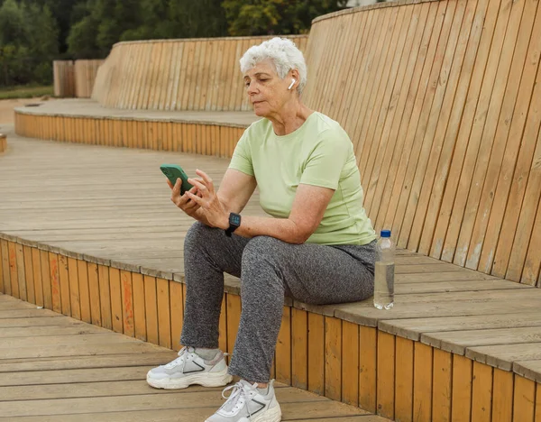 An elderly happy retired woman after a run sits on a wooden bench and sends a text message. Modern technology, sports, lifestyle and modern seniors concept.
