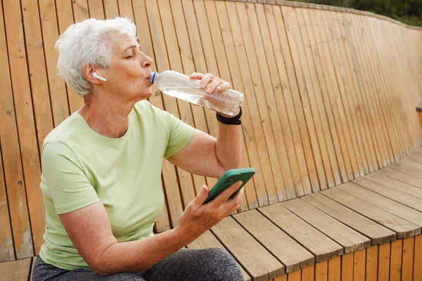 An elderly happy pensioner woman after a run sits on a wooden bench and drinks water. Modern technology, sports, lifestyle and modern seniors concept.