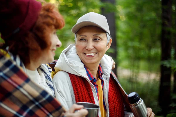 Two elderly woman friends walk in the forest, pour coffee from a thermos, have a great time together, an elderly happy couple.