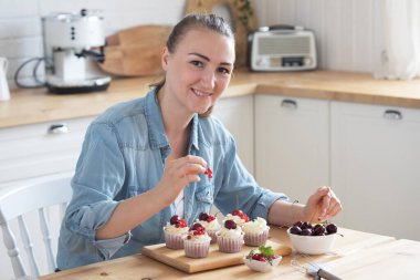 A young woman decorates muffins with white cream and berries in her kitchen. Food and people concept.