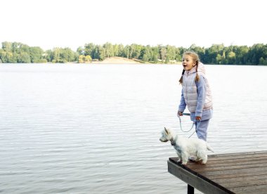 A seven-year-old girl with a small white dog stands on a bridge near the lake and looks at the water. Friendship with pets. Happy childhood.