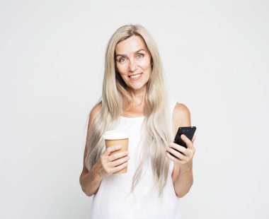 Elderly woman with long hair holding smartphone and takeaway coffee over light grey color background