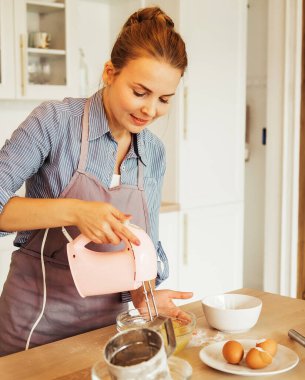 Young blonde woman uses mixer and whips cream for cake, stands in modern kitchen