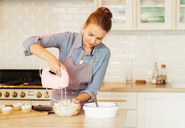 Young blonde woman uses mixer and whips cream for cake, stands in modern kitchen