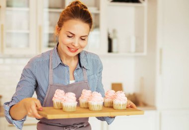 Work in confectionery workshop. Handmade cupcakes. Smiling pretty young woman pastry chef, demonstrating tasty fresh cupcales with pink cream to camera.