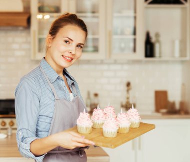 Work in confectionery workshop. Handmade cupcakes. Smiling pretty young woman pastry chef, demonstrating tasty fresh cupcales with pink cream to camera.
