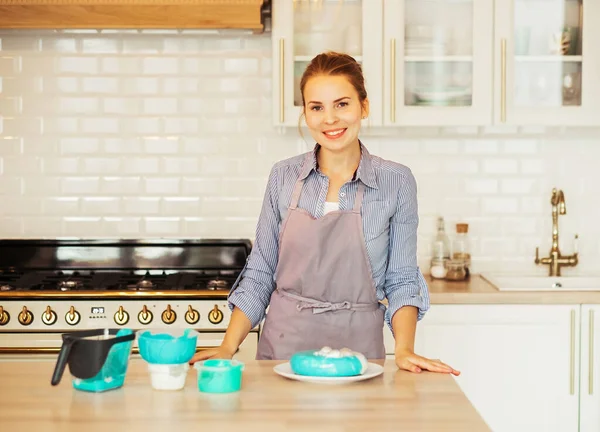 Young blonde woman decorating a cake with blue icing, modern kitchen, freelancing and hobby concept