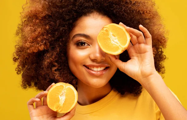 lifestyle, food, diet and people concept: Photo of young dark skin female hide eye half slice orange wear t-shirt isolated on yellow color background