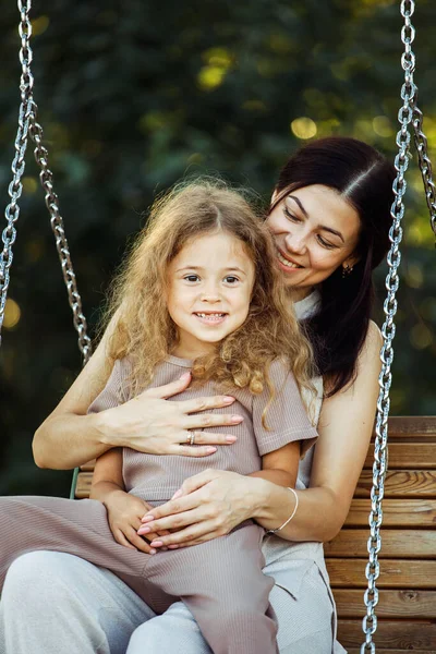 Mom and daughter swing on a swing. Caucasian young happy woman and her little girl have fun on the playground.