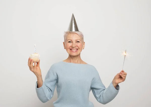 党と高齢者の概念 幸せな中年女性身に着けています円錐形の帽子祝いの誕生日とともにカップケーキとキャンドルと輝き以上グレーの背景 — ストック写真