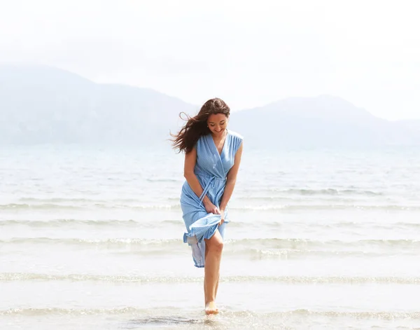 青いドレスを着た若いブルネットの女性がビーチを裸足で歩き 水の中で足をぶら下げています ライフスタイルコンセプト — ストック写真
