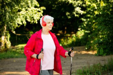 Sporty elderly woman trains endurance while Nordic walking in nature. Old female listening music in headphones.