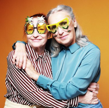Two elderly women friends wearing party glasses hugging on yellow background. Lifestyle, friendship and old people concept.
