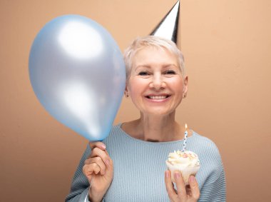 Portrait of her she nice-looking cheery gray-haired lady wearing party hat holding blue balloon and cupcake with candle over beige background.