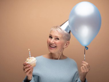 Portrait of her she nice-looking cheery gray-haired lady wearing party hat holding blue balloon and cupcake with candle over beige background.