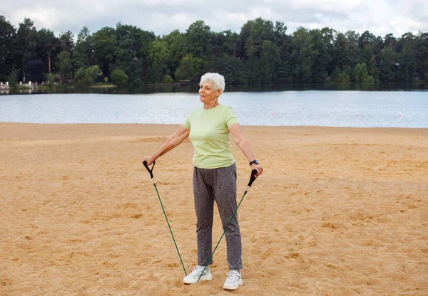 Elderly woman wear sportswear doing exercises outdoor on the beach in the morning using resistance rubber bands. Healthy lifestyle, active retired life and sporty time.